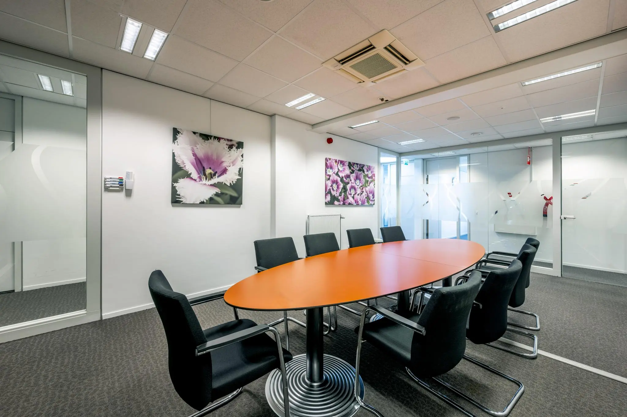 Vergaderruimte aan de Jan Tooropstraat met een oranje ovale tafel, zwarte stoelen en kleurrijke bloemenfoto’s aan de muur.